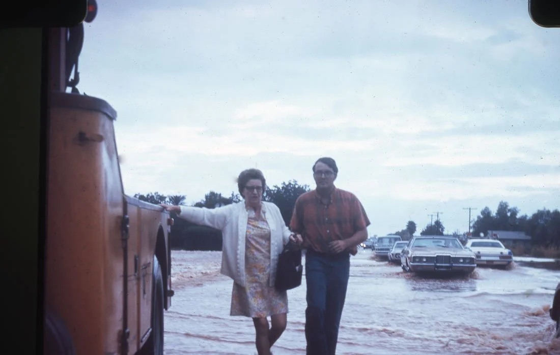 A woman and a man walking through a muddy parking lot, with several cars parked in the background. The woman is wearing a light-colored dress and white jacket, and the man is wearing glasses and a red plaid shirt.