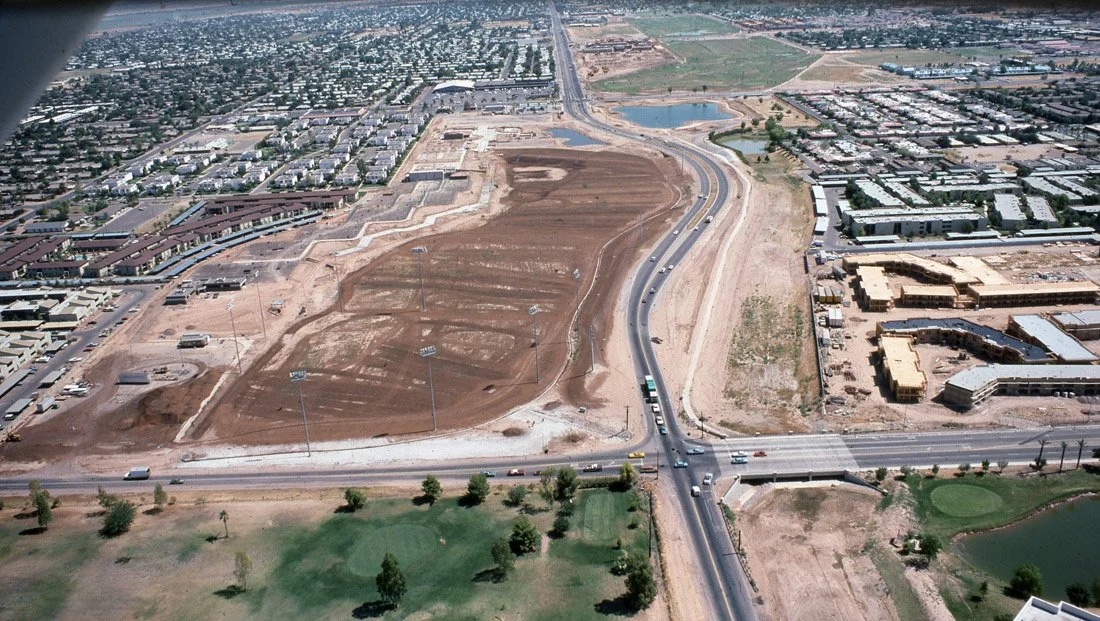 Aerial view of a construction site next to a busy highway intersection in a city, with ongoing land development and surrounding buildings, parks, and water bodies.