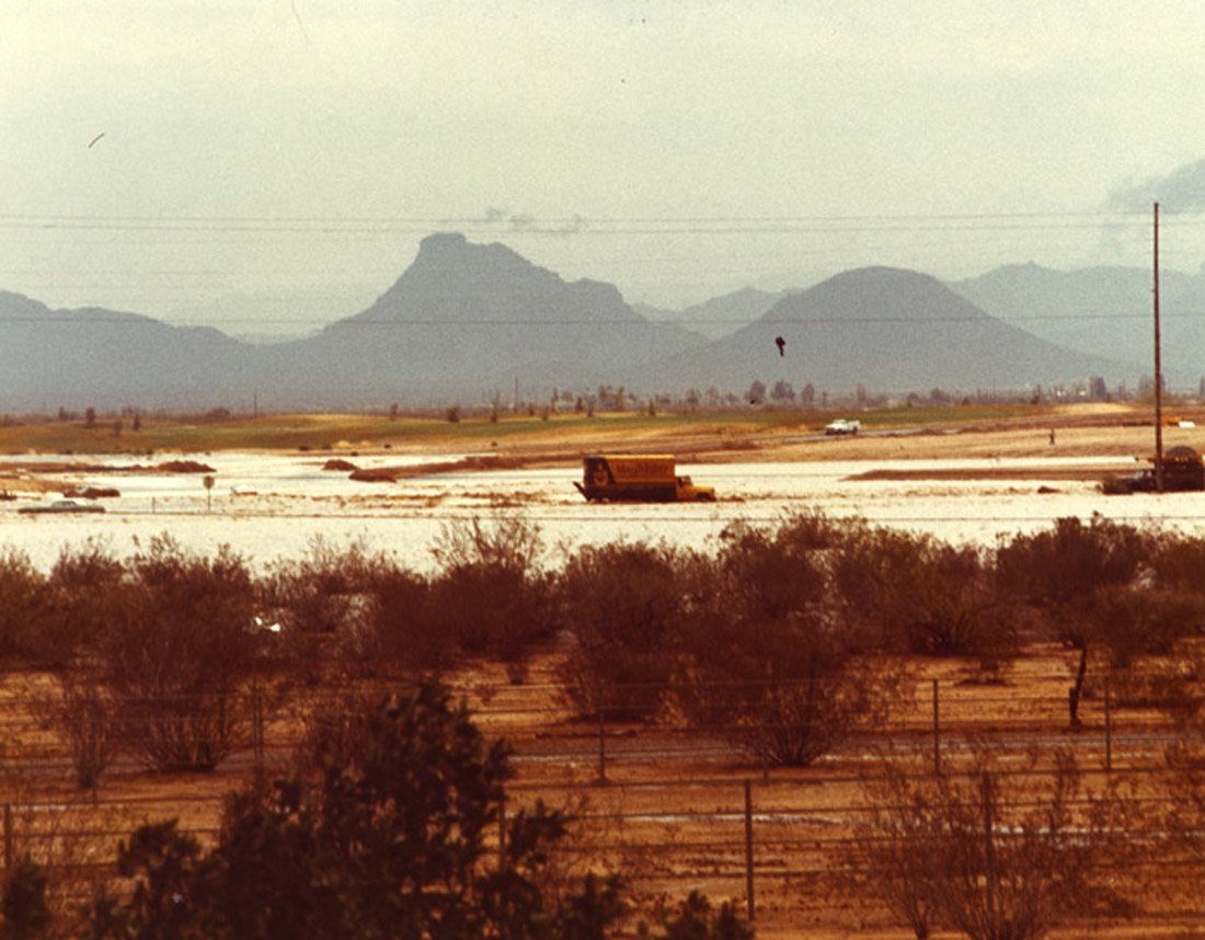 Desert landscape with sparse vegetation, water or salt flats, distant mountains, and a clear sky.