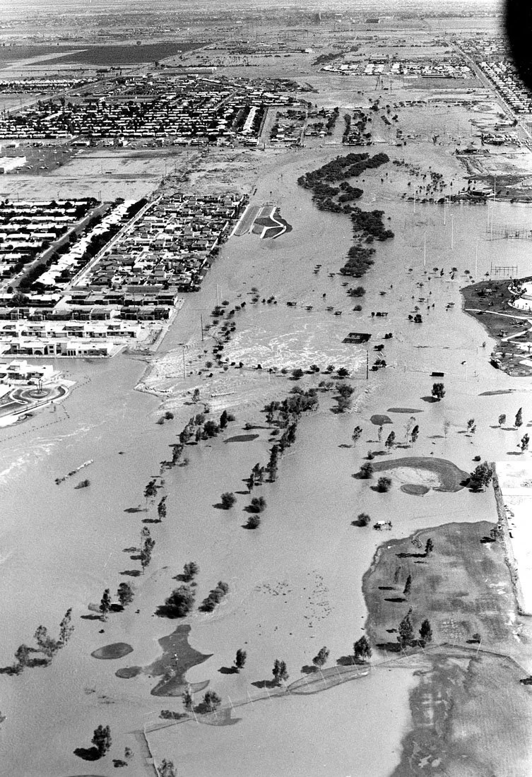 Aerial view of a flood covering a city park and surrounding neighborhoods with floodwaters, trees, and some buildings partially submerged.