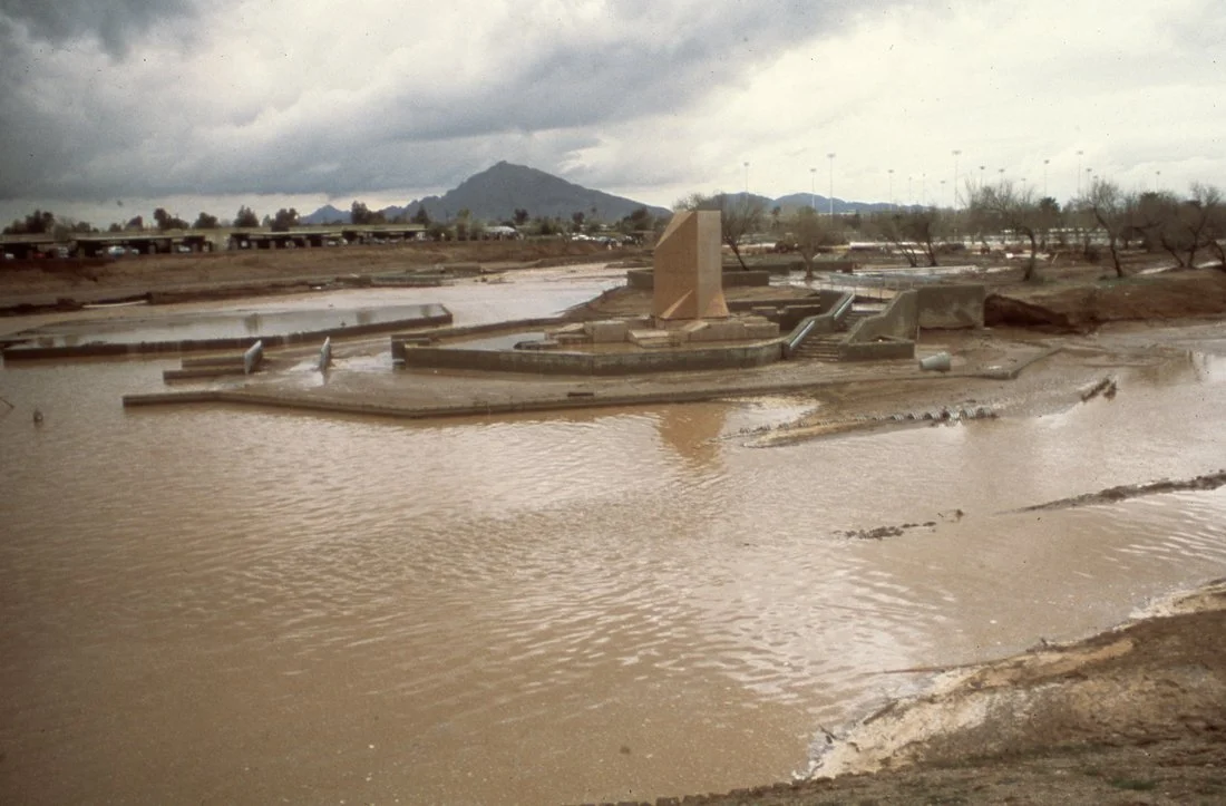 Flooded area with concrete structures and a small water feature, on a cloudy day with mountains in the background.