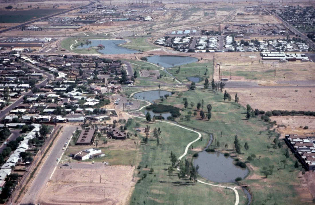 Aerial view of a park with a winding creek, small lakes, grassy areas, trees, and surrounding residential neighborhoods and empty lots.