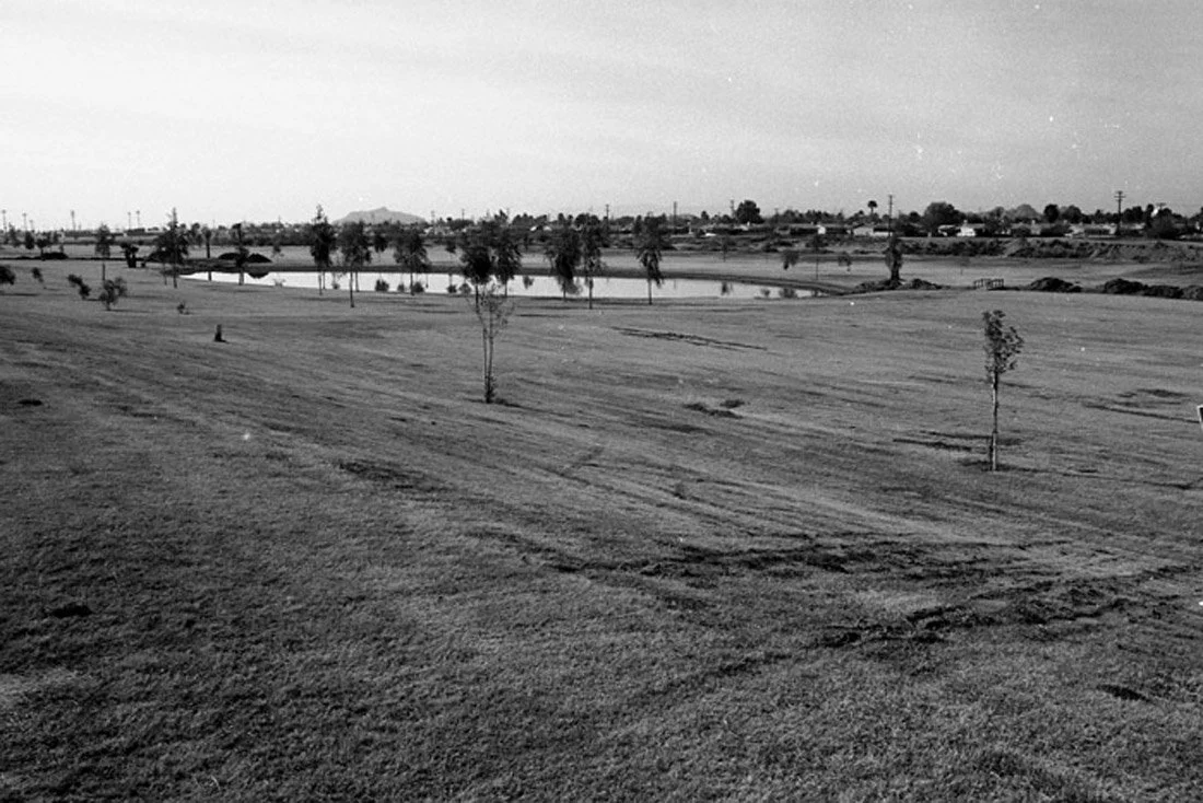 A black and white photo of a golf course with a few small trees, a pond, and distant buildings and hills in the background.