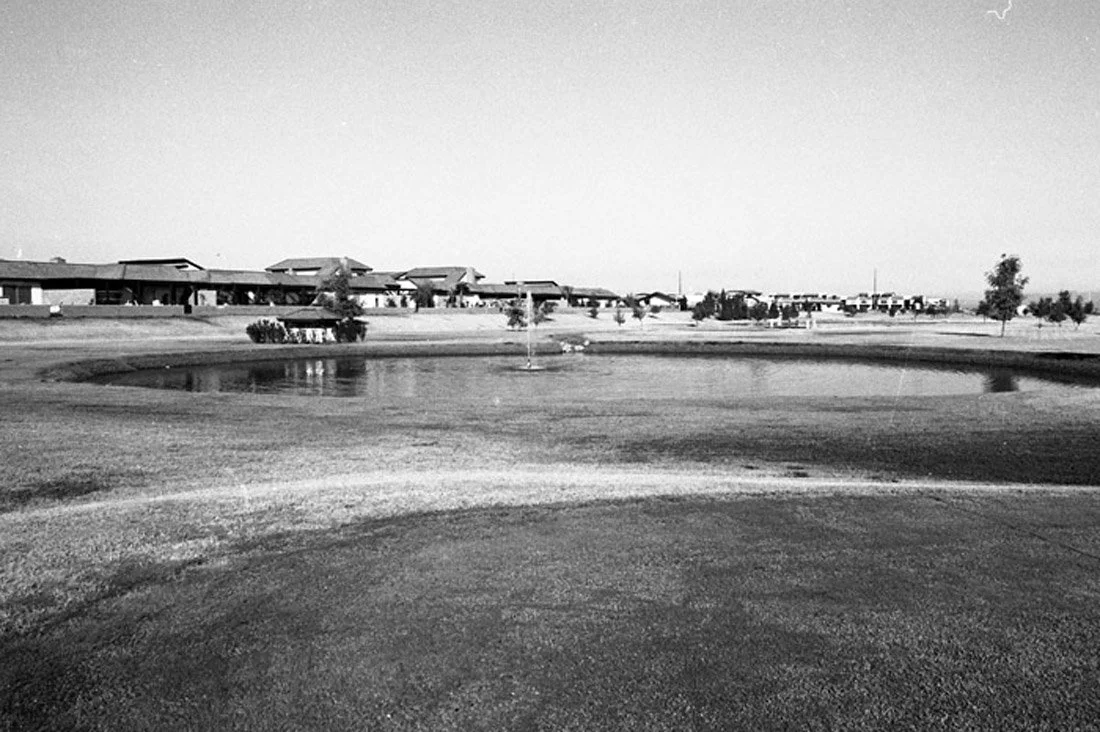 A black and white photograph of a residential neighborhood with single-story houses in the distance, a small pond, and a fountain in the foreground, with a clear sky above.