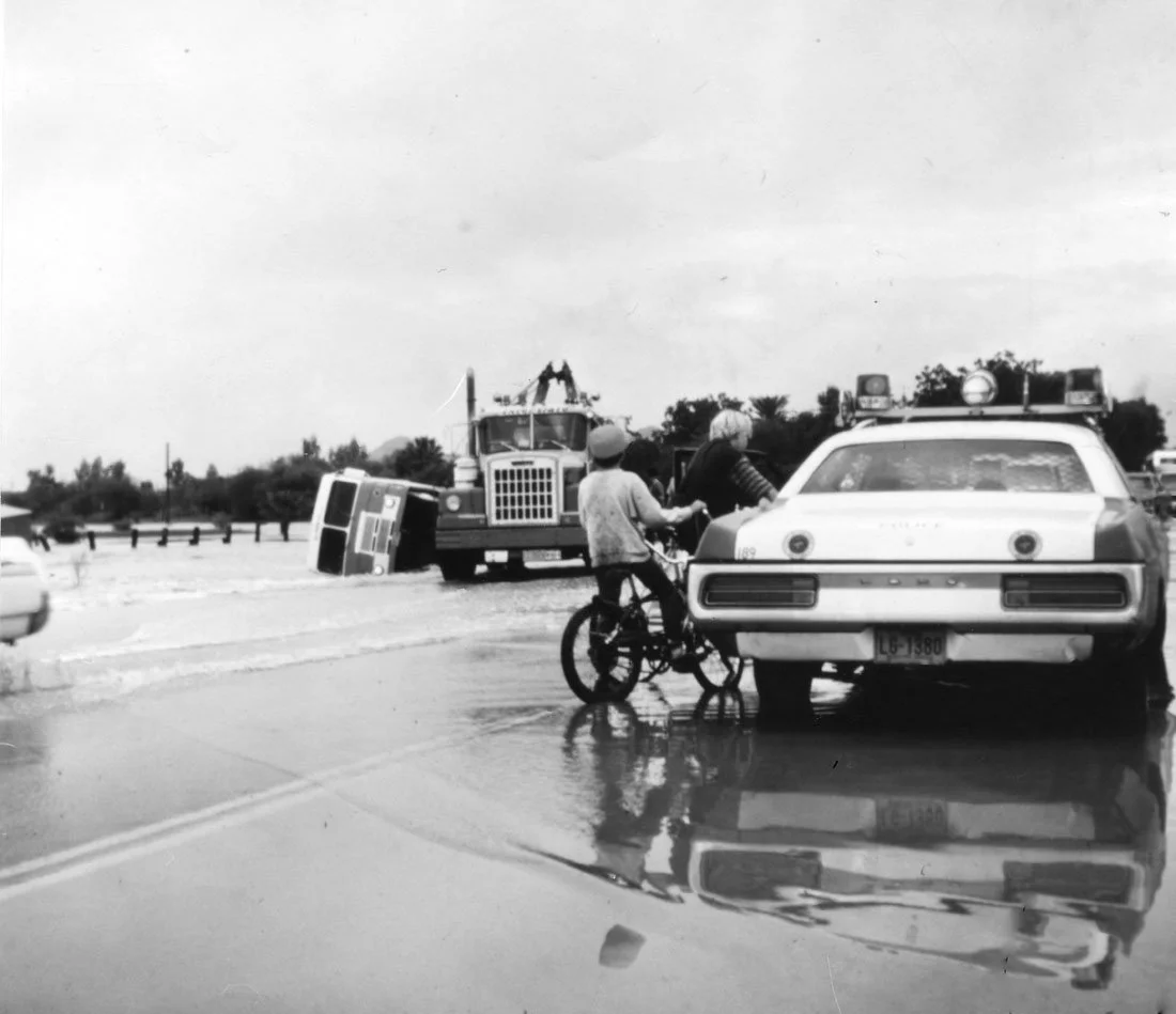 A person on a bicycle holding onto a car, with an overturned truck and flooding in the background.