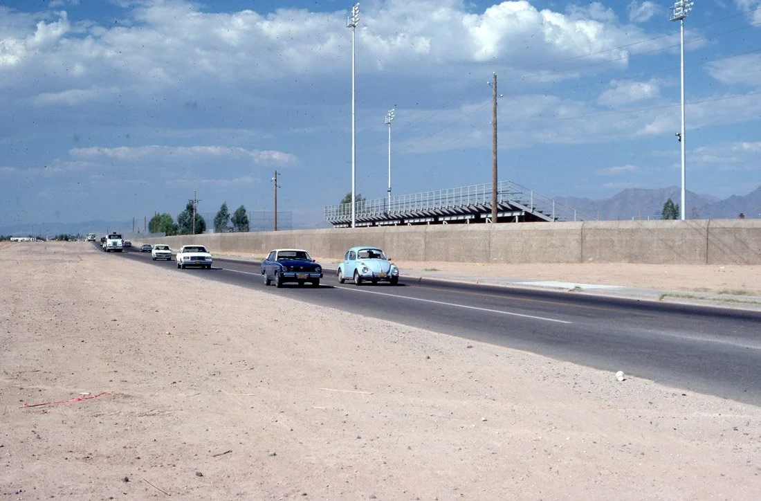 A highway with a few vintage cars driving on it, bordered by a concrete wall, with empty bleachers and light poles, under a partly cloudy sky, with distant mountains.