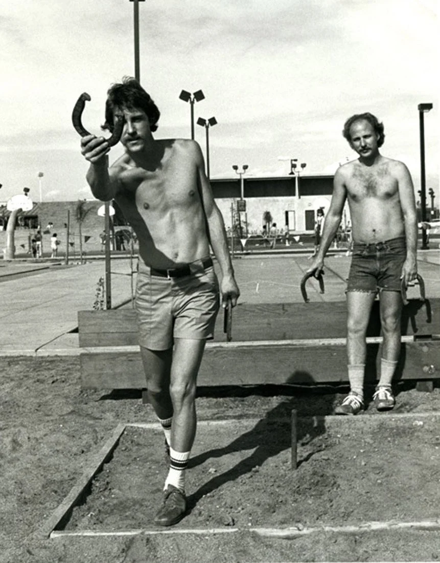 Two shirtless young men with mustaches on a playground or skate park. One is standing on the ground, holding a hockey stick and wearing shorts, socks, and sneakers. The other is standing on a wooden platform, holding a hockey stick, wearing shorts, s