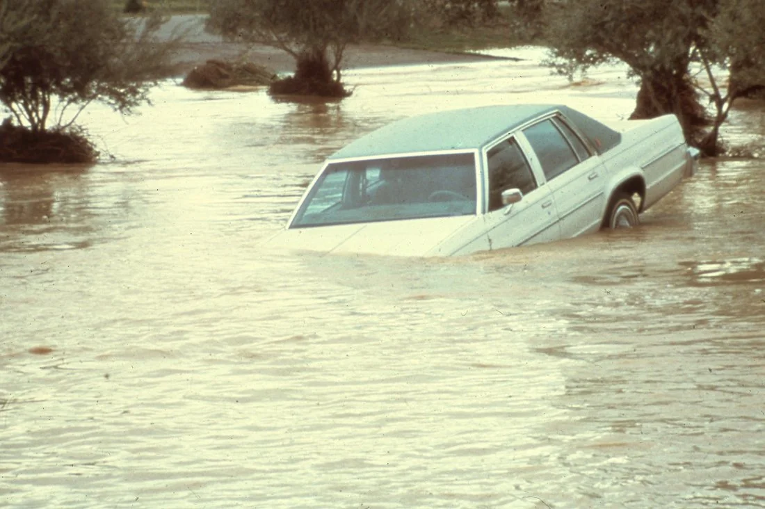 A silver sedan partially submerged in muddy floodwaters with trees around it.