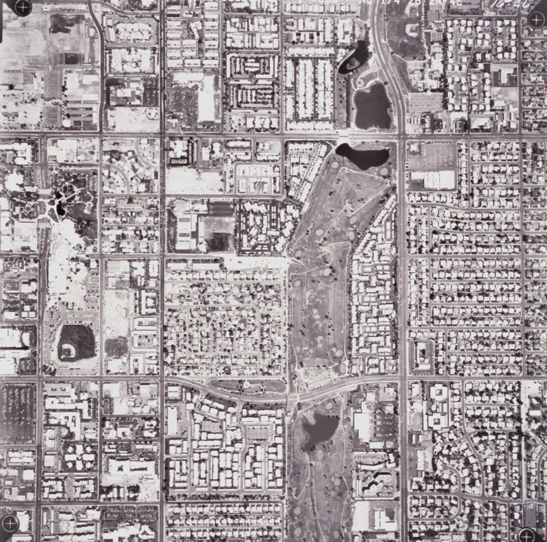 Aerial black-and-white photograph of an urban city grid with streets, buildings, residential neighborhoods, parks, and lakes.