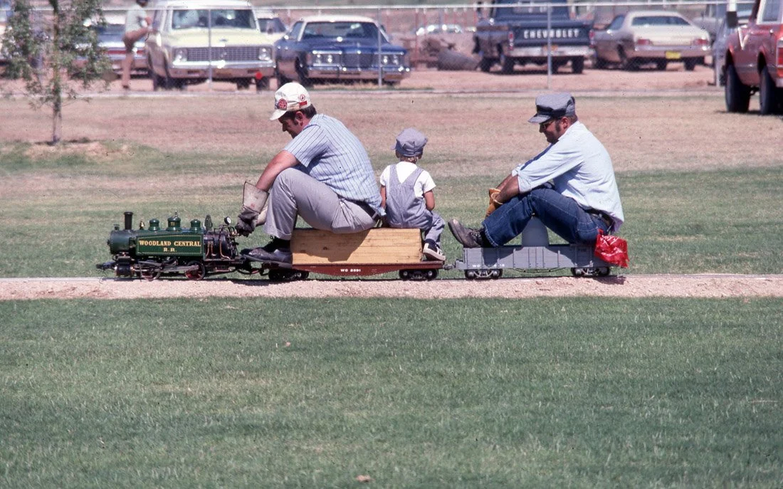 Three people riding on a miniature train on a dirt path in a grassy park, with cars parked in the background.