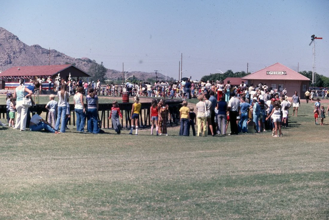 Crowd of people watching a carnival or fairground game with a house and mountains in the background.