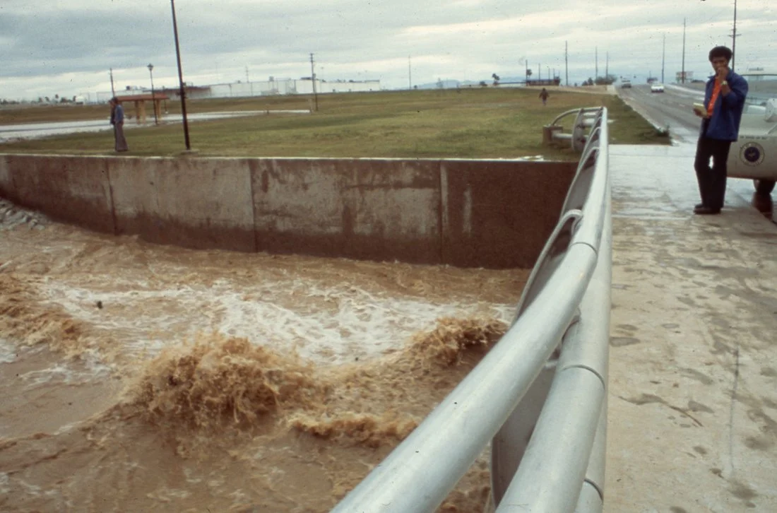 Flooded sidewalk next to a grassy park, with muddy water spilling over a concrete wall and rushing onto the sidewalk; a man is standing on the sidewalk near a parked car, holding a cellphone and wearing a blue jacket.