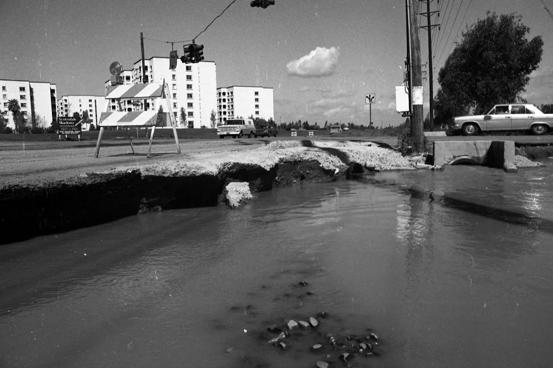 A broken street with a large pothole filled with water, next to a construction barricade. In the background, there are apartment buildings, a traffic light, cars, and power lines, with a cloudy sky overhead.