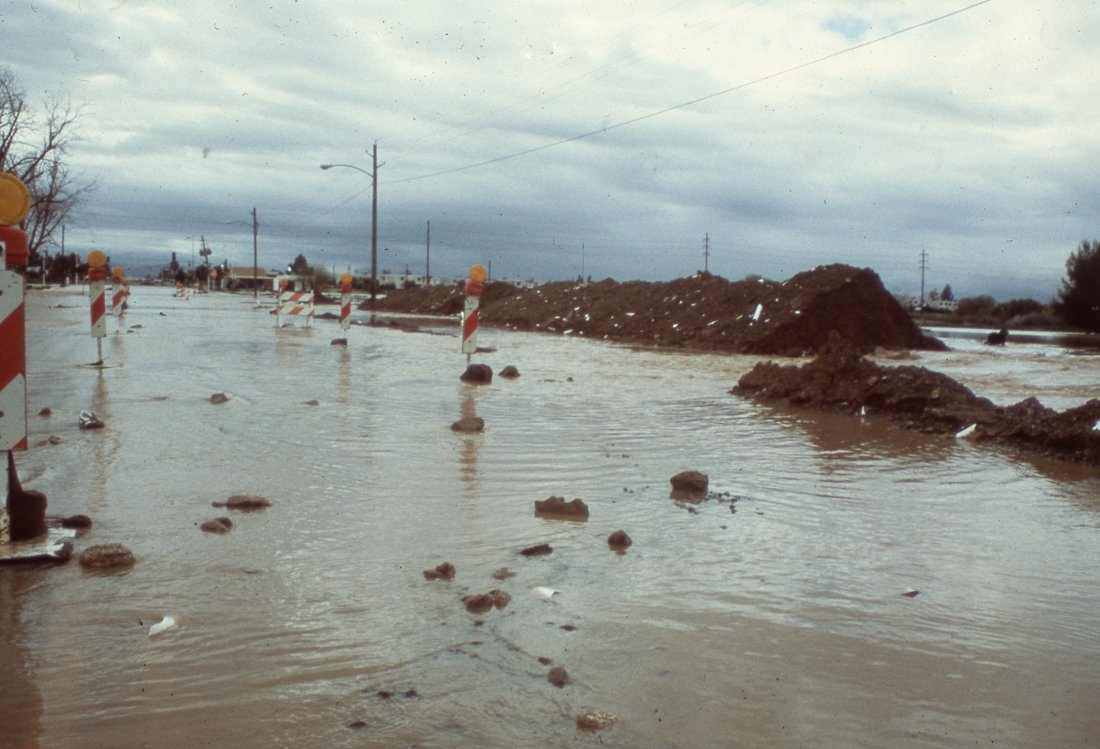 Flooded road with water covering the surface, construction barriers, and large dirt piles on either side, under a cloudy sky.
