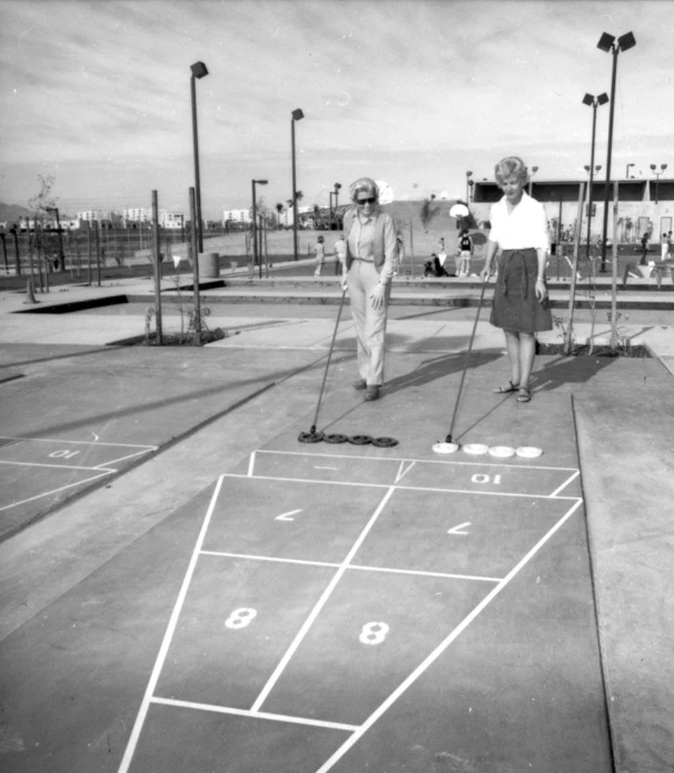 Two women are playing shuffleboard outdoors on a concrete court, with one holding a shuffleboard disc and the other preparing to shoot. The court has painted lines and numbers, and there are other people and a park area in the background.