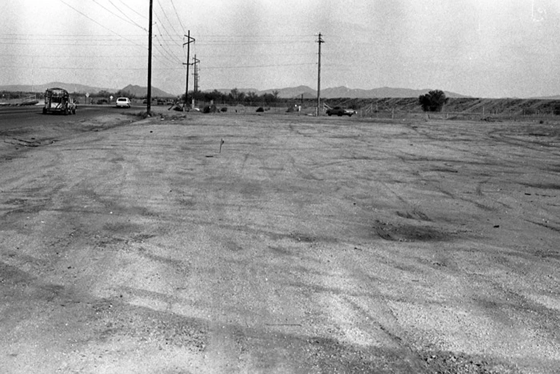Deserted dirt lot with power lines, a bus, cars, and distant mountains in the background.