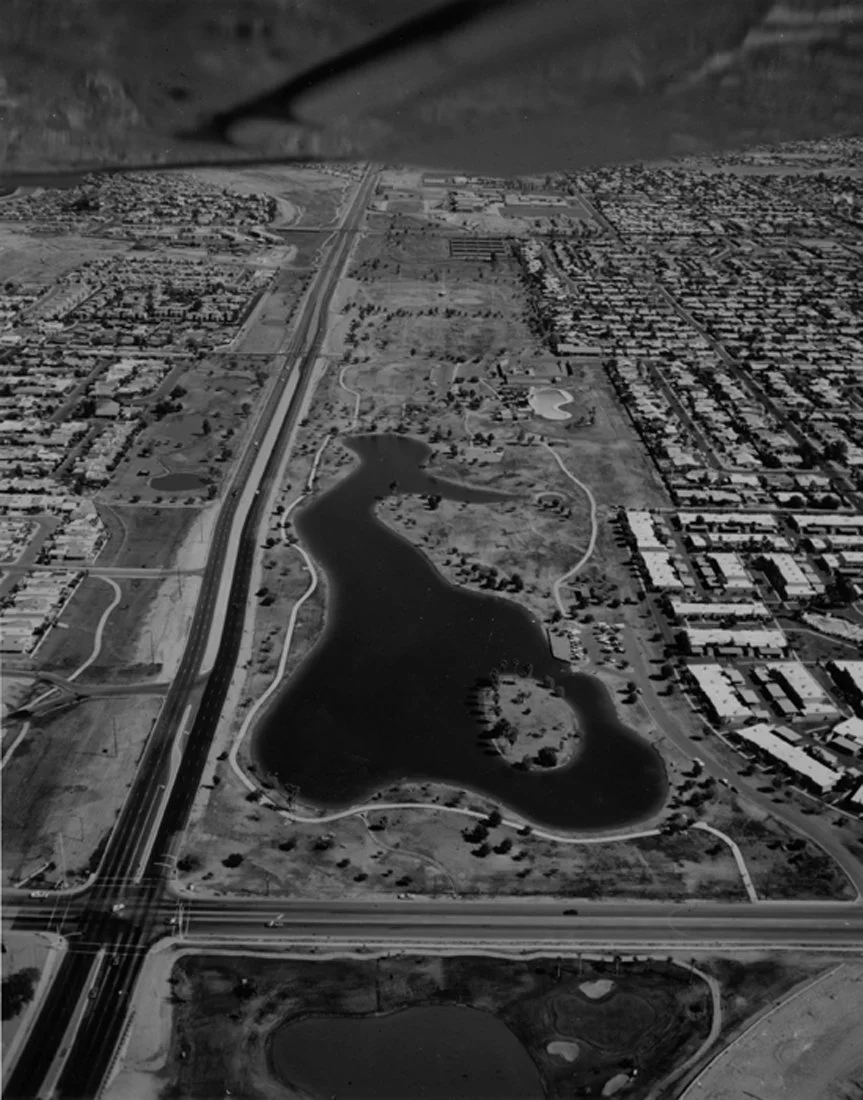 Aerial view of a city park with a large lake, walking paths, residential neighborhoods, and highways.