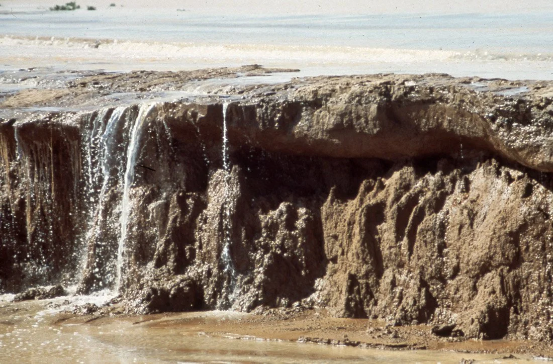 A rocky shoreline with water flowing over the edge, creating a small waterfall, on a sandy beach.