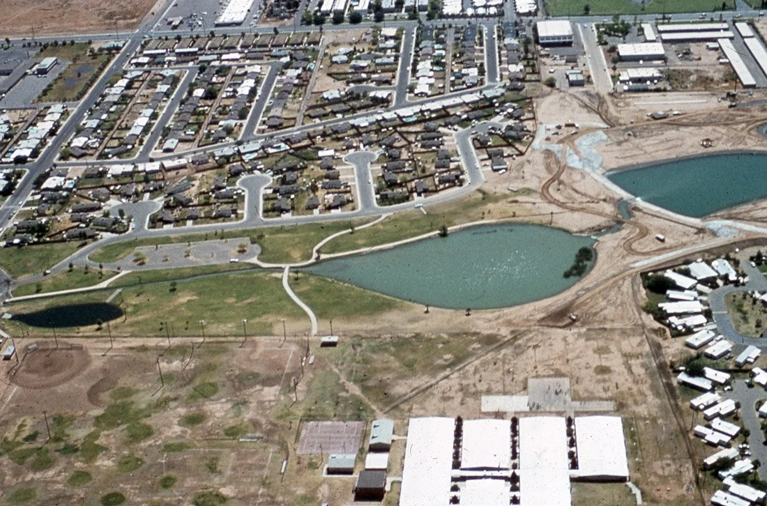 Aerial view of a park with lakes, walking paths, and open grassy areas, surrounded by residential and industrial buildings.