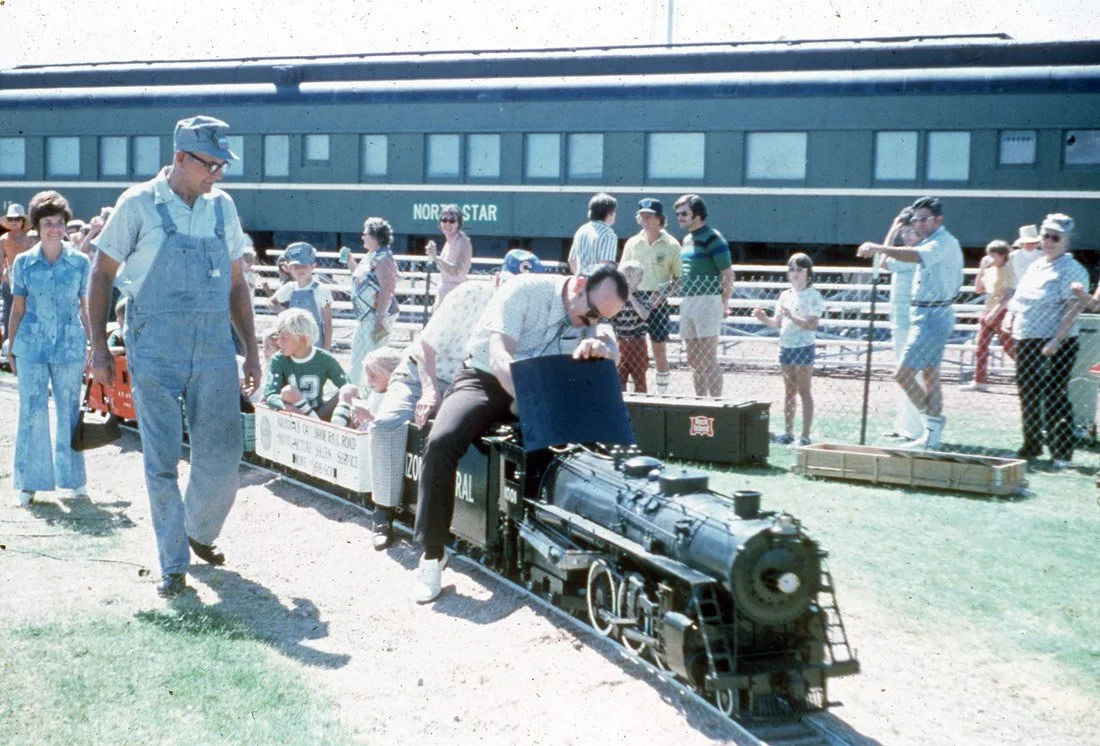 People riding a small toy train with a large train car in the background at an outdoor event in a grassy area.