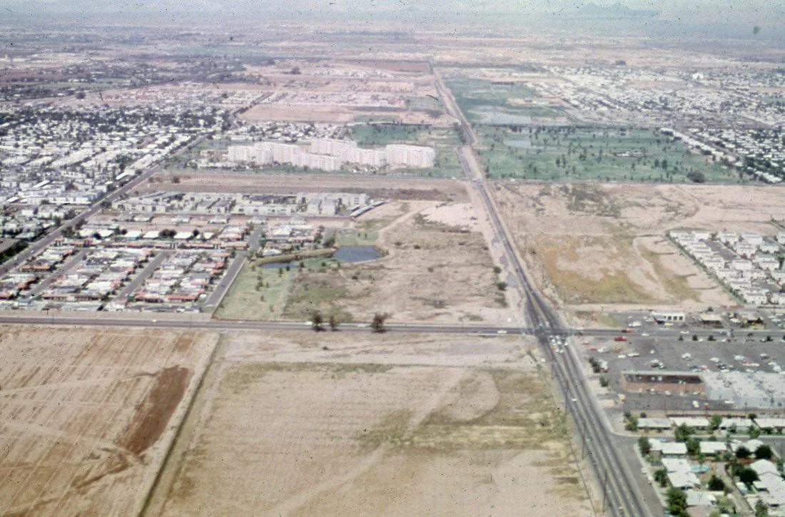 Aerial view of a city landscape with a mix of residential and commercial buildings, roads, open land, fields, and a small pond.