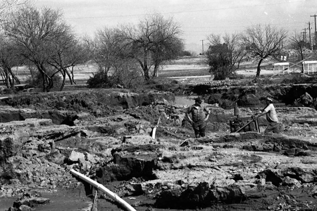 Black and white photo of two men working in an archaeological dig site with exposed rocks and dirt. Bare trees are in the background.