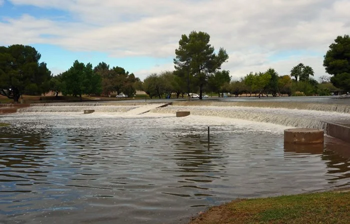 A river or pond with water cascading over a small dam, surrounded by trees and a cloudy sky.