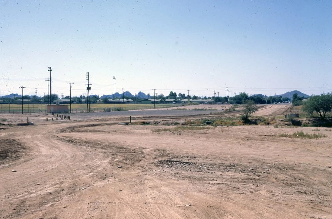 A dirt road in a rural area with power lines and sparse vegetation under a clear blue sky.