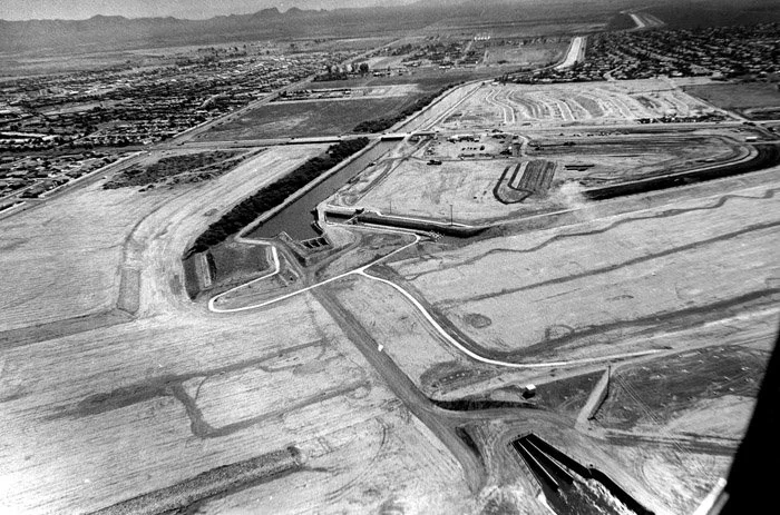 Aerial black and white photo of a construction site with roads, a canal, and land development in a flat area, possibly near a city or town.