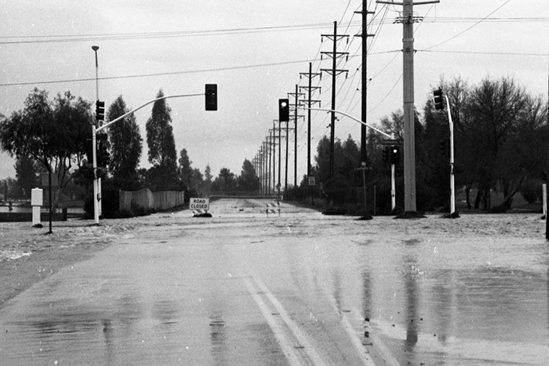 Flooded road with traffic lights, power lines, and a road closed sign, with trees in the background.