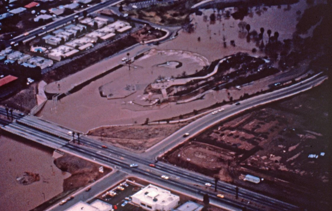 Aerial view of a flood-damaged area with water covering streets and land, featuring roads, a bridge, and partly submerged buildings.