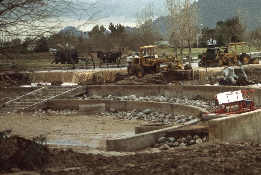 Construction site with bulldozers and a partially built concrete structure in a field with mountains and trees in the background.