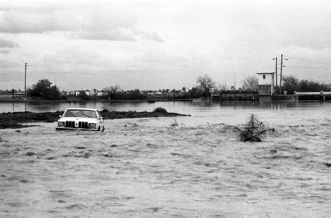 A black-and-white photo of a flooded area with a partially submerged car and a small tree in the water.