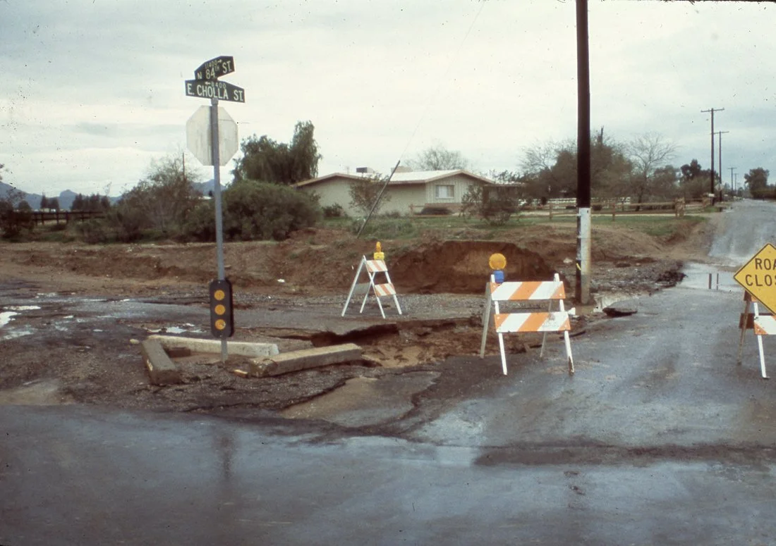 Road construction with barriers, warning signs, and a large pothole or damaged road surface at an intersection of N 84th Street and E Cholla Street in a suburban area.
