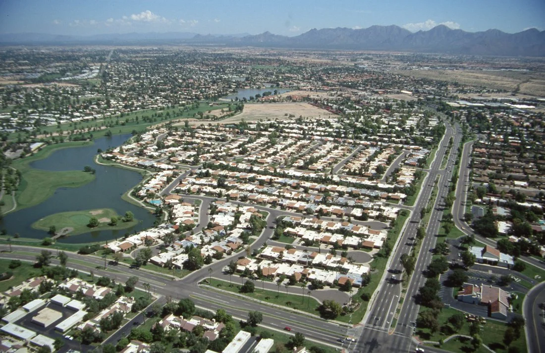 Aerial view of a suburban city with numerous houses, roads, a golf course, a river, and mountains in the background.