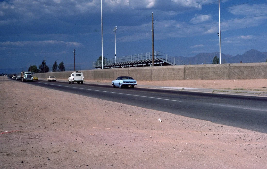 A highway with cars driving in both directions, set against a backdrop of mountains and a cloudy sky, with a baseball lying on the sandy shoulder of the road.