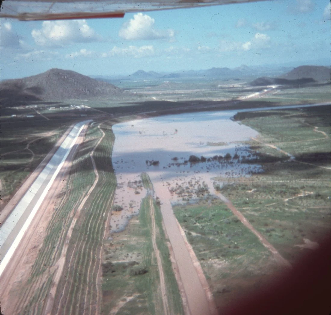 Aerial view of a landscape with a river, agricultural fields, and mountain ranges in the distance, taken from an airplane window.