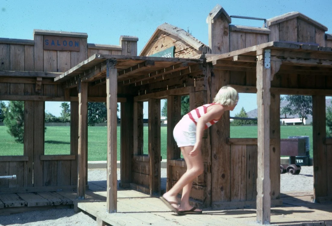 A person with light blond hair wearing a red and white striped tank top and white shorts leaning forward in front of a rustic wooden playhouse or structure, outdoors on a sunny day with green grass and trees in the background.