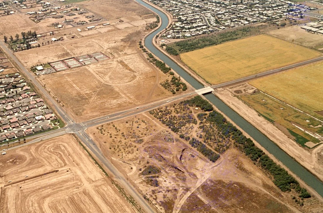 Aerial view of a landscape with residential neighborhoods, agricultural fields, a canal, and undeveloped land with dirt paths and sparse vegetation.