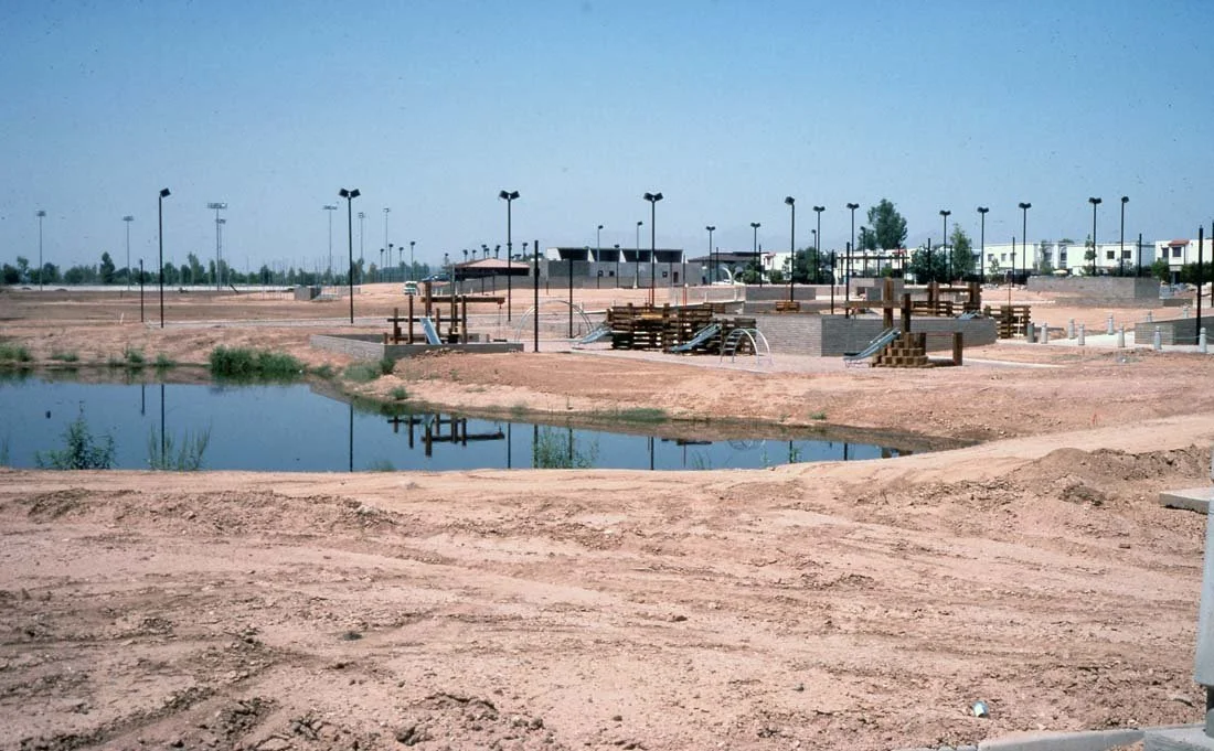 An under-construction park or public space with a small pond, construction materials, and several light poles on a clear day.