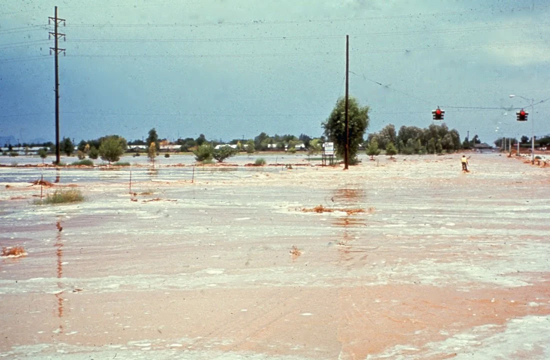 An empty, dry, and cracked landscape with sparse trees, power lines, and traffic lights hanging over an intersection under a blue sky.