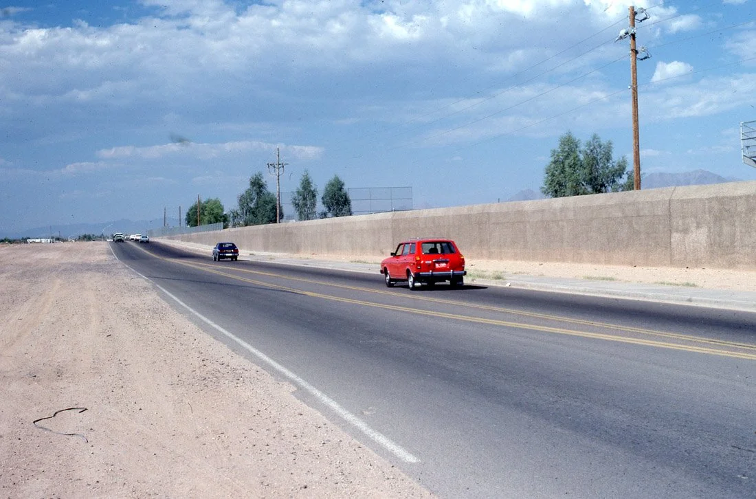 A straight road with a red van and some other cars, desert landscape, utility poles, and cloudy sky.