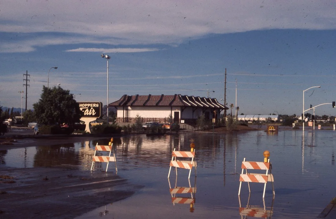 A flooded parking lot with orange and white barricades partially submerged in water, a building in the background, and a sign reading "Hoppers Air" visible.