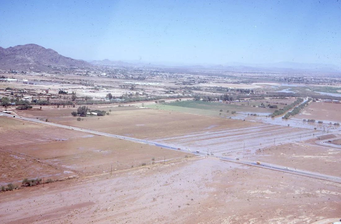 Aerial view of a flat, arid landscape with fields, roads, and some water flooding parts of the land in a rural area with mountains in the background.