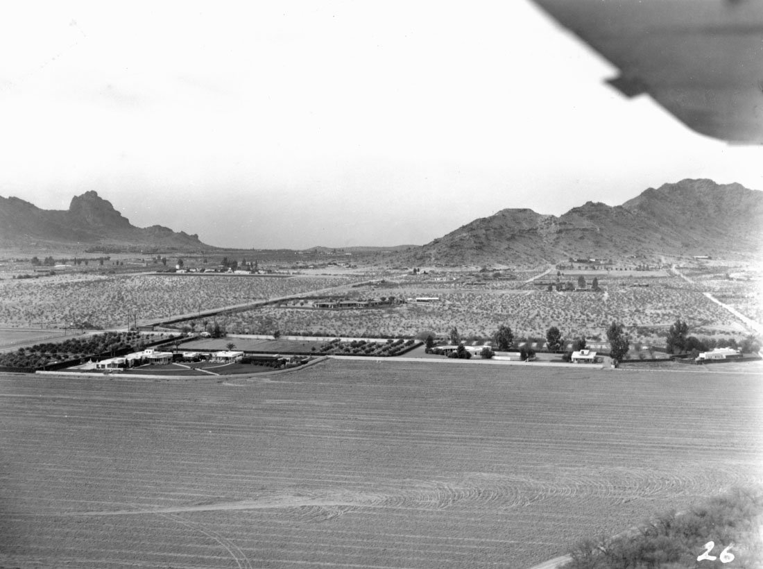 Black and white aerial photograph of rural farmland with a few buildings, dirt roads, trees, and distant mountains under a cloudy sky.