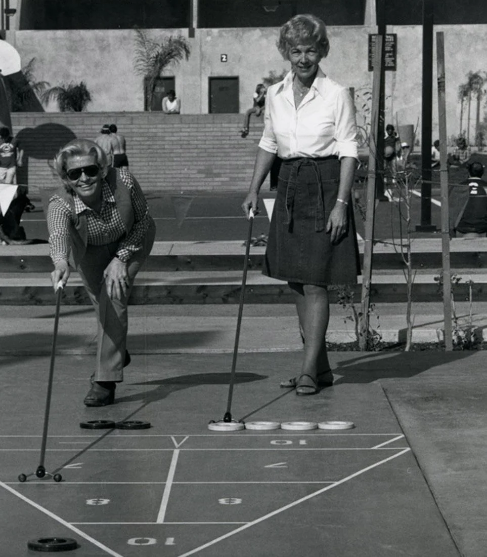 Two women playing shuffleboard outdoors, one with sunglasses and checkered shirt leaning forward, the other in a white blouse and dark skirt standing upright, both holding cues and aiming at the shuffleboard court.