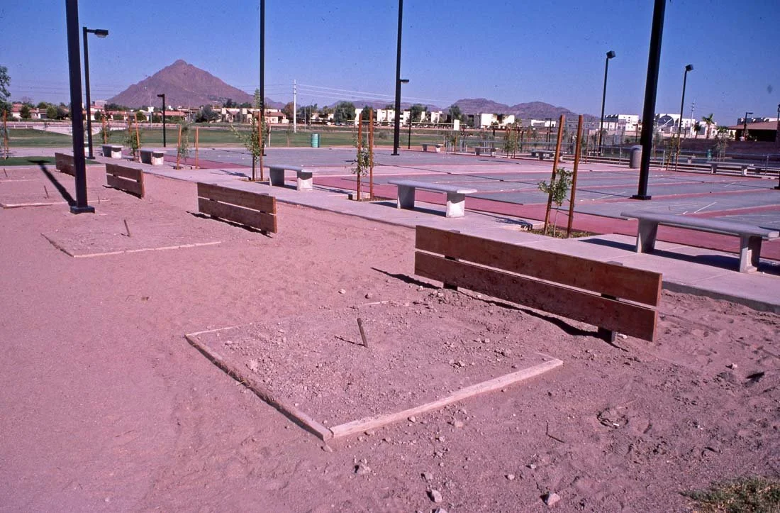 Empty outdoor tennis court with benches, young trees, and mountains in the background on a clear day.