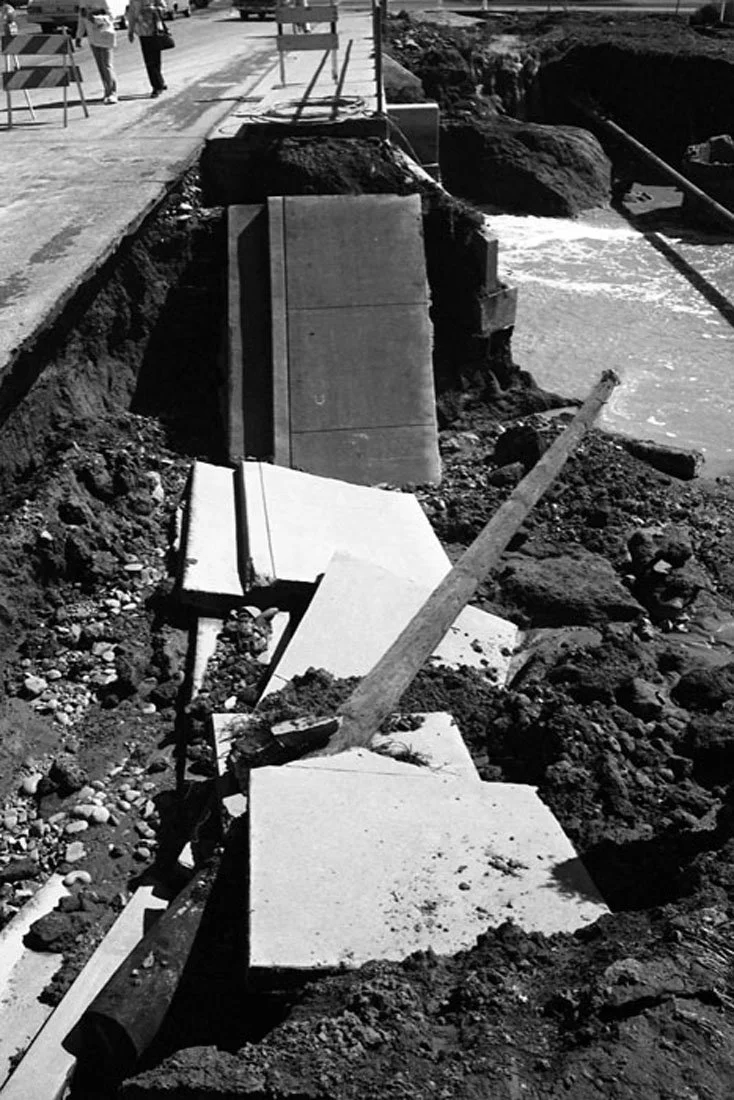 A damaged sidewalk with broken concrete slabs and a leaning wooden plank, adjacent to an open excavation site with muddy water, and traffic cones and barriers in the background.