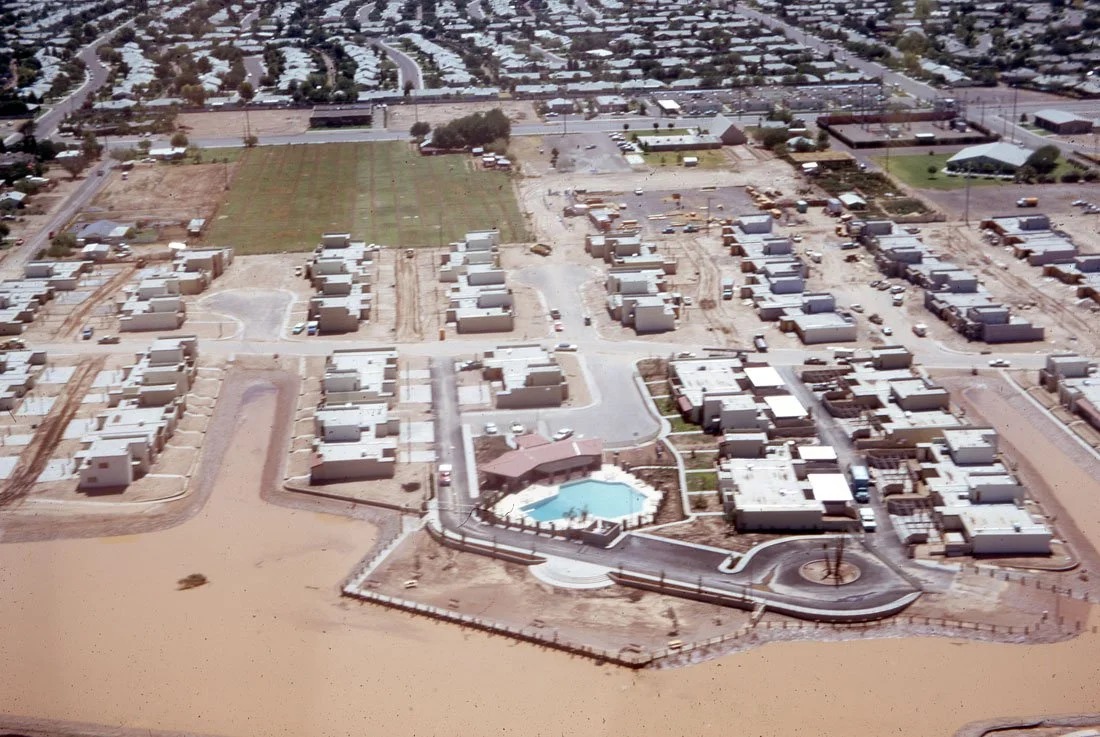 Aerial view of a residential area with newly constructed houses, a swimming pool, streets, and a large water body in the foreground.