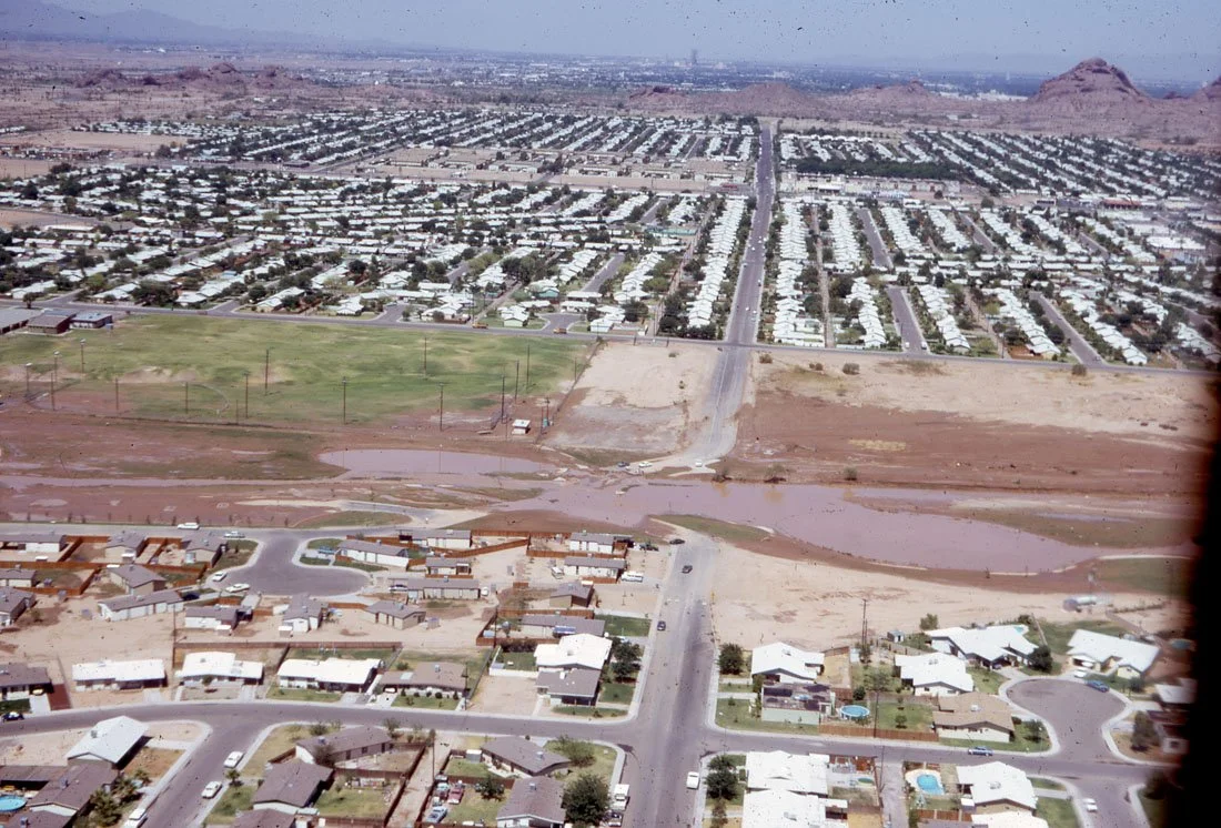 Aerial view of a suburban neighborhood and a large residential area with a park, baseball field, a flooded area, and hills in the background.